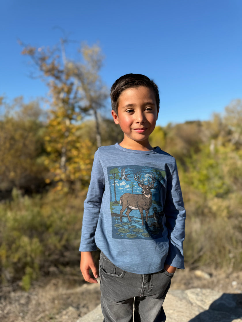 A boy wearing a blue long-sleeve graphic tee featuring a deer in a woodland scene.