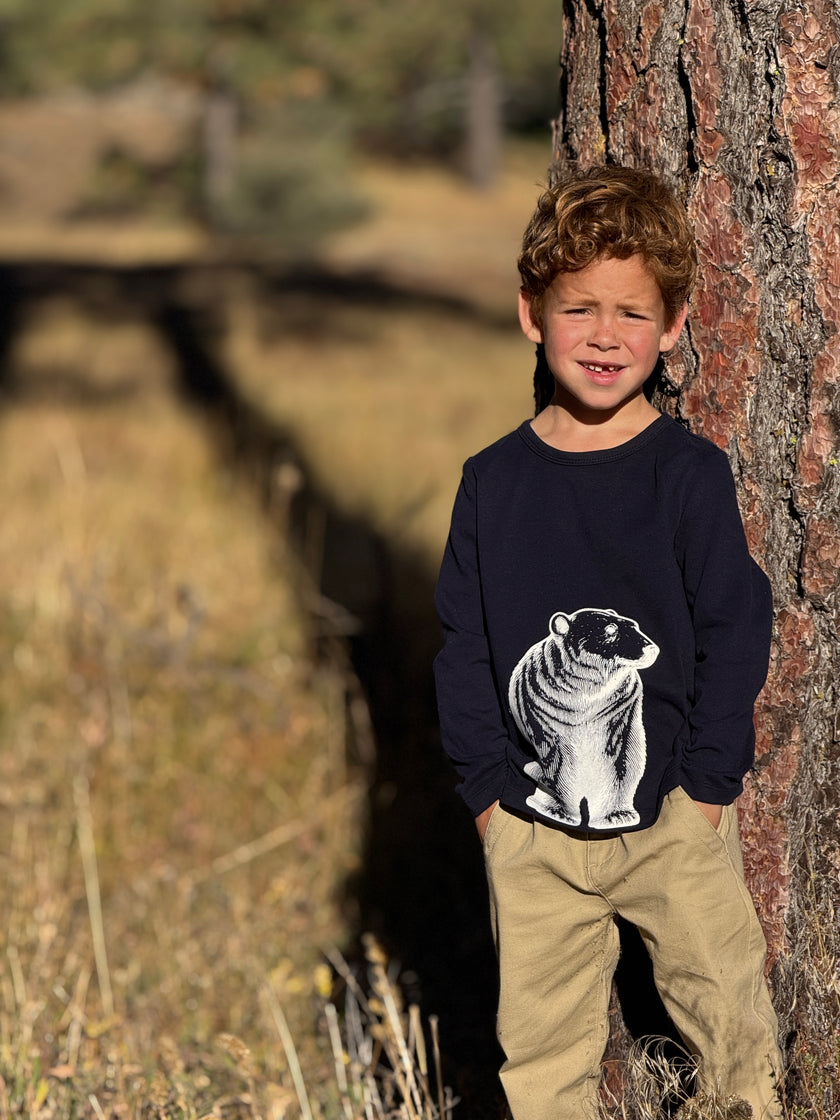 A young boy with curly light brown hair stands outdoors leaning against a large tree trunk.