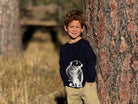 A young boy with curly light brown hair stands outdoors leaning against a large tree trunk, wearing a navy blue long-sleeve shirt with a white graphic of a seated animal and khaki pants.