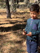 A young boy wearing a teal and navy striped long-sleeve henley shirt and navy pants stands in a sunny, dry outdoor forest setting, looking down at a small brown object in his hand.