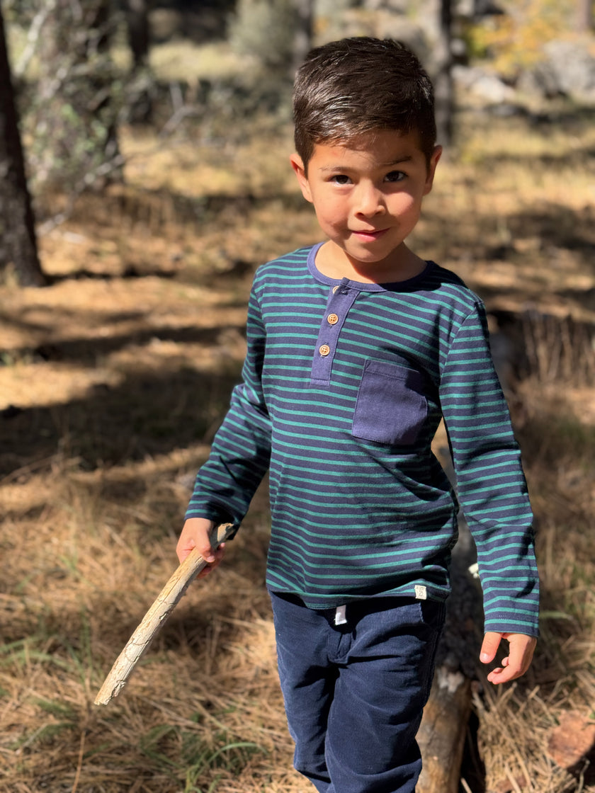 A young boy wearing a navy and teal striped long-sleeve henley shirt with a chest pocket, standing outdoors in a wooded area and holding a small stick.