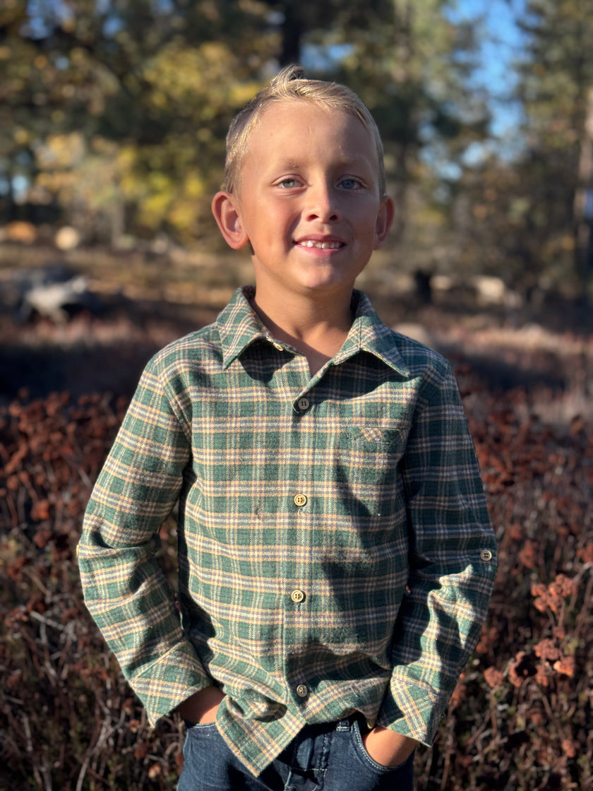 A young boy with blond hair stands outdoors wearing a green and beige plaid long-sleeve button-up shirt and blue jeans, with a blurred natural background of trees and brush.