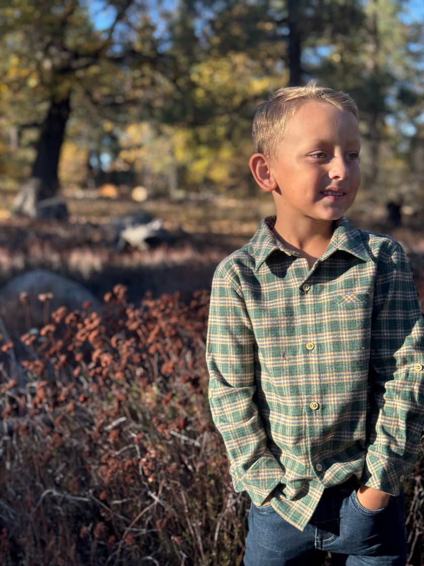 A young boy wearing a green and beige plaid long-sleeve button-up shirt and blue jeans, standing outdoors in a wooded area with autumn foliage.
