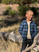 A young boy wearing a blue and green plaid flannel button-up shirt over a cream t-shirt and dark jeans, standing outdoors in a grassy, wooded area.
