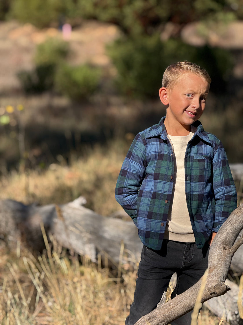 A young boy wearing a blue and green plaid flannel button-up shirt over a cream t-shirt and dark jeans, standing outdoors in a grassy, wooded area.