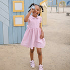 A young child wearing a pink striped dress and white sandals, standing outdoors in front of a blue and yellow wall while holding up a Mushie kaleidoscope to one eye.