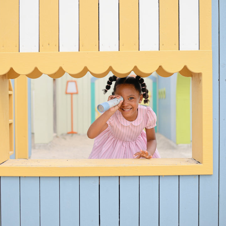 A young child with pigtails wearing a pink collared dress, looking through a Mushie kaleidoscope while peering through a yellow-framed cutout in a blue and white striped playhouse wall.