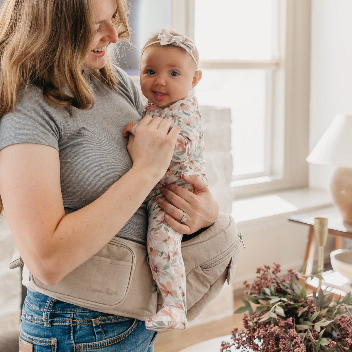 Woman holding a baby in a sling in a bright room with flowers on a table.