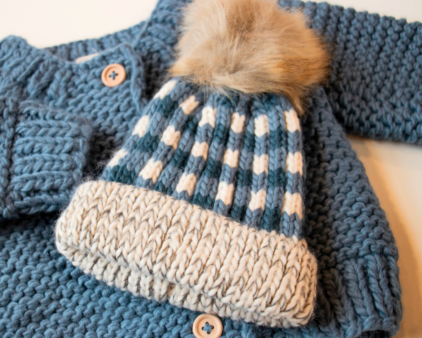Blue and white checkerded beanie with a beige puff ball on top on a blue knit sweater from Huggalugs