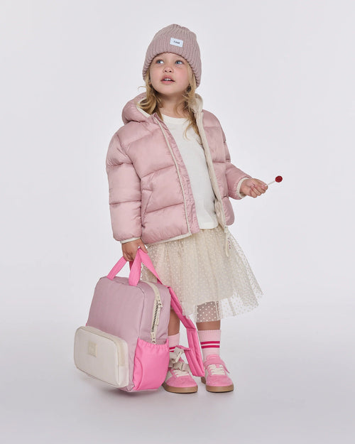 A young girl stands against a light gray studio background wearing a pale pink puffer jacket, cream tulle skirt, pink beanie, and pink sneakers, while holding a pink and cream backpack.
