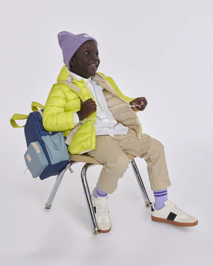 A child model wearing a bright lime green puffer jacket, beige pants, white sneakers, a purple beanie, and purple socks, sitting on a metal chair with a blue and light blue backpack against a light gray studio background.