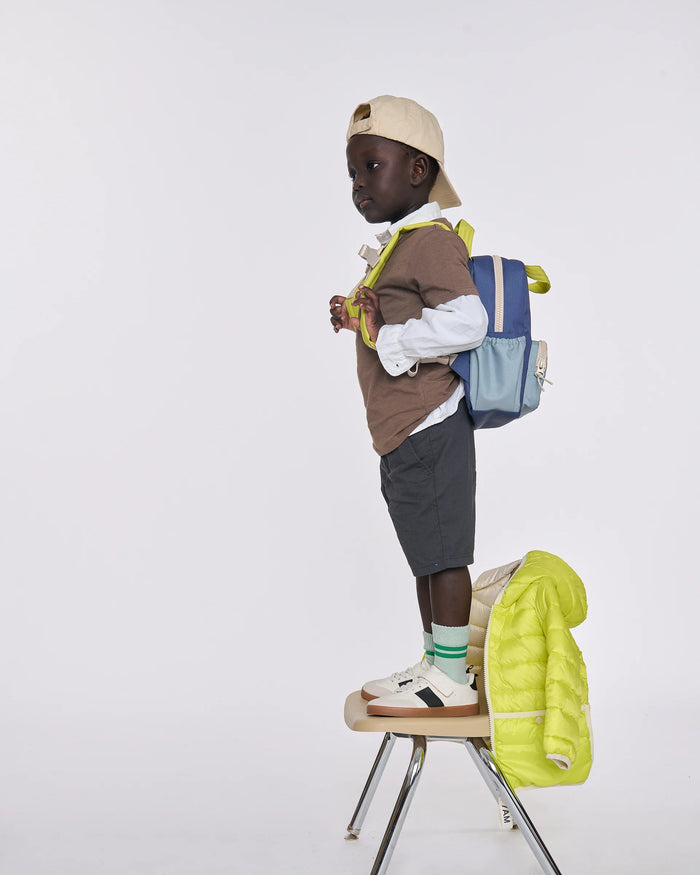 A child model stands on a chair in profile wearing a beige cap, brown vest, white shirt, dark shorts, white sneakers, a blue and gray backpack, and a bright yellow puffer jacket draped over the chair, against a light gray studio background.
