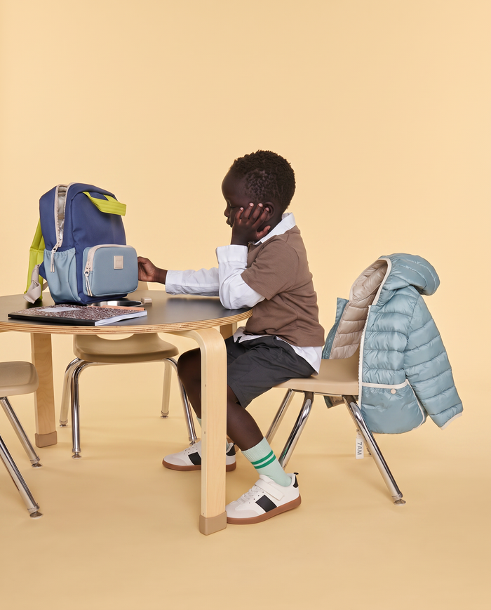 A child sits at a small wooden table wearing a brown vest, white shirt, dark shorts, striped socks, and white sneakers. A blue backpack with lime green accents sits on the table, and a light blue puffer jacket is draped over the chair.