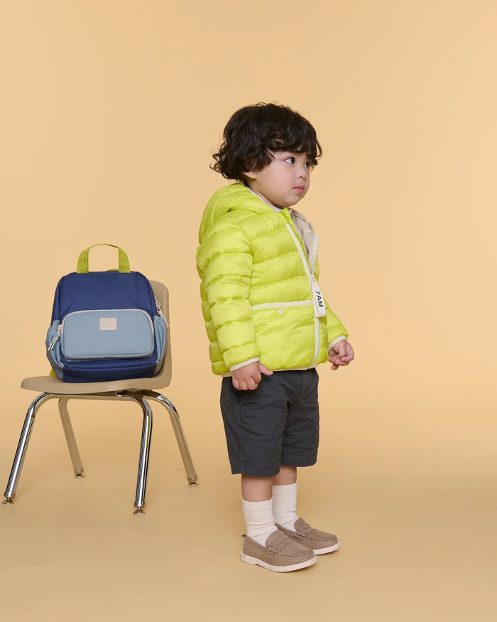 A toddler stands in profile wearing a bright lime green puffer jacket, dark gray shorts, white socks, and tan slip-on shoes, posed next to a blue and light blue backpack on a chair against a solid beige studio background.