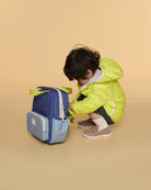 A child in a bright lime green puffer jacket crouches beside a blue and light blue backpack on a solid beige studio background.