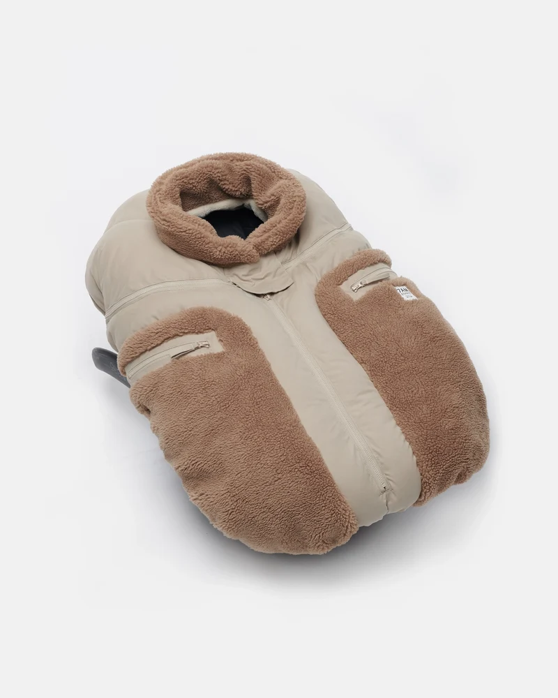 Benji Applique Car Seat Cocoon by 7AM Enfant featuring a close-up of the light taupe or oat-colored weatherproof outer shell, brown sherpa fleece trim around the collar and zippered side pocket, and a front zipper detail.
