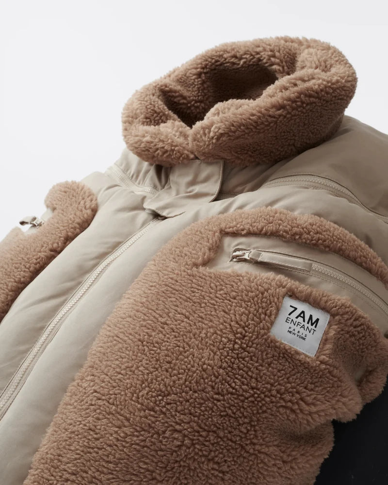 Benji Applique Car Seat Cocoon by 7AM Enfant featuring a close-up of the light taupe or oat-colored weatherproof outer shell, brown sherpa fleece trim around the collar and zippered side pocket, and a branded 7AM Enfant patch.