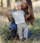 A smiling woman crouches outdoors in a grassy field while holding and supporting a toddler boy. The boy is wearing a light cream henley shirt and beige-and-white vertically striped pants.
