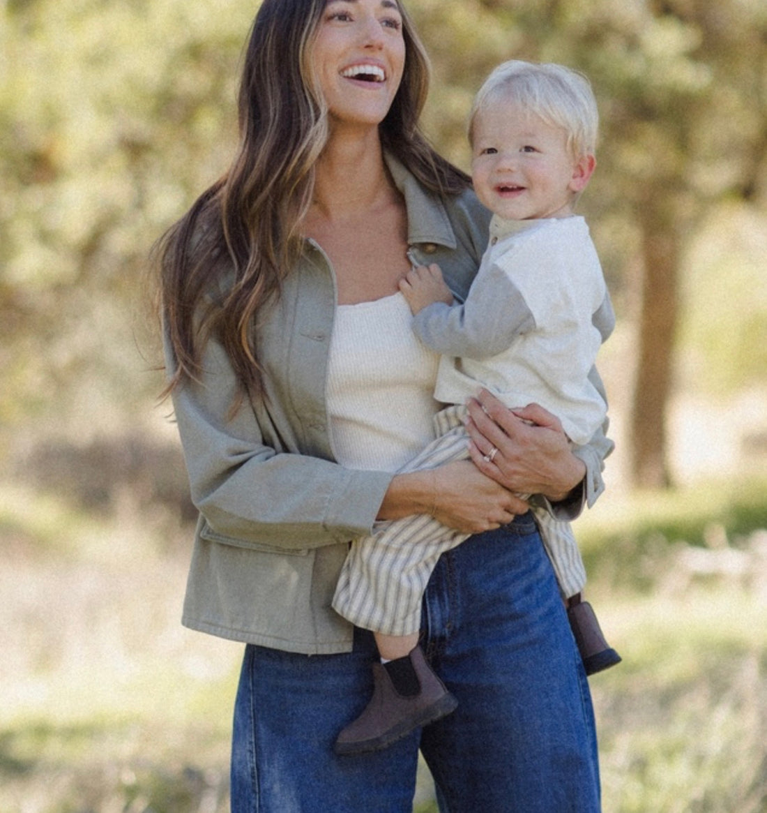 A woman outdoors holding a smiling toddler. The woman wears an olive green utility jacket over a white top and blue jeans, while the child wears a white long-sleeve shirt and light striped pants.