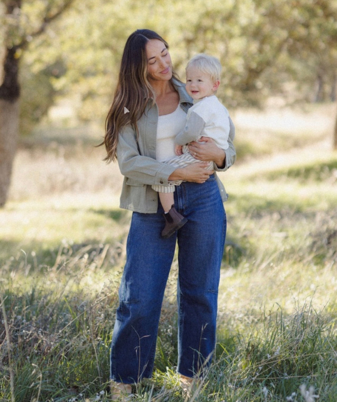 A woman standing in a grassy outdoor field holds a baby. She is wearing a light sage green button-up shirt over a white top and wide-leg blue jeans.