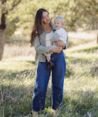 A woman standing in a grassy outdoor field holds a baby. She is wearing a light sage green button-up shirt over a white top and wide-leg blue jeans.