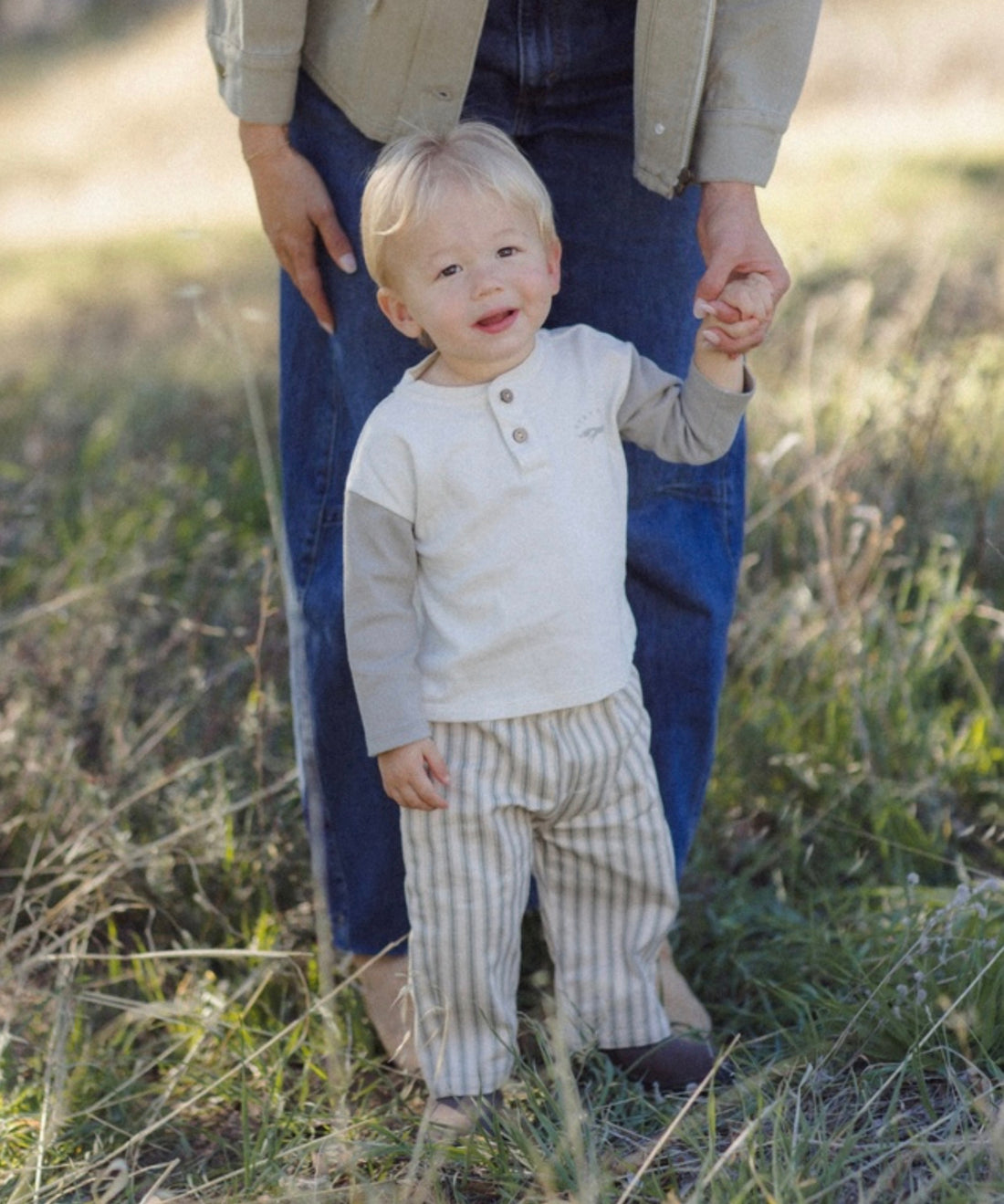 A toddler standing outdoors in a grassy field holding an adult's hand. The child is wearing a cream long-sleeve henley top and cream-and-beige vertically striped pants.