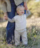 A toddler wearing a cream henley shirt with gray sleeves and beige-and-white vertically striped pants, standing outdoors in a grassy field while holding an adult's hand.