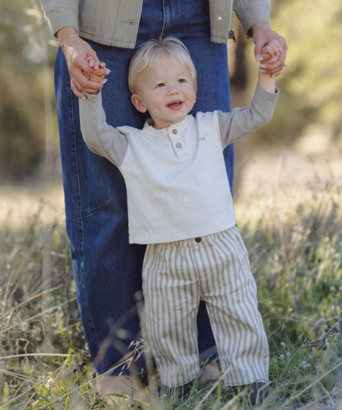 A toddler standing outdoors in a grassy field, holding hands with an adult. The child is wearing a light beige henley shirt with long gray sleeves and beige-and-white vertically striped pants.
