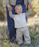A toddler standing outdoors in a grassy field, holding hands with an adult. The child is wearing a light beige henley shirt with long gray sleeves and beige-and-white vertically striped pants.