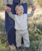 A toddler standing outdoors in a grassy field holding hands with an adult wearing blue jeans and a beige jacket. The child is wearing a light beige henley shirt with gray sleeves and beige-and-white vertically striped pants.