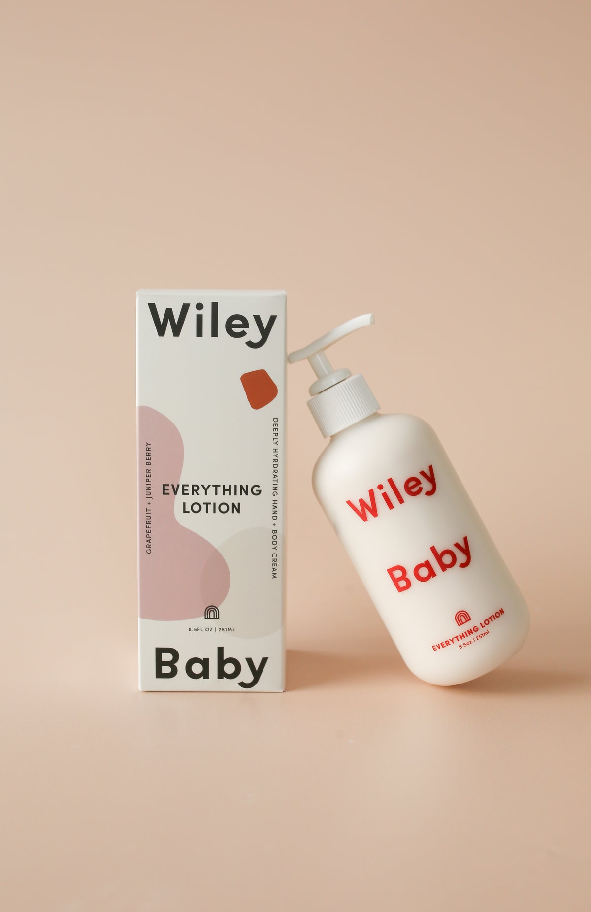 Wiley Baby Everything Lotion bottle and packaging box on a solid pale peach background. The bottle is white with red text, and the box is white with black text and a pink abstract shape.