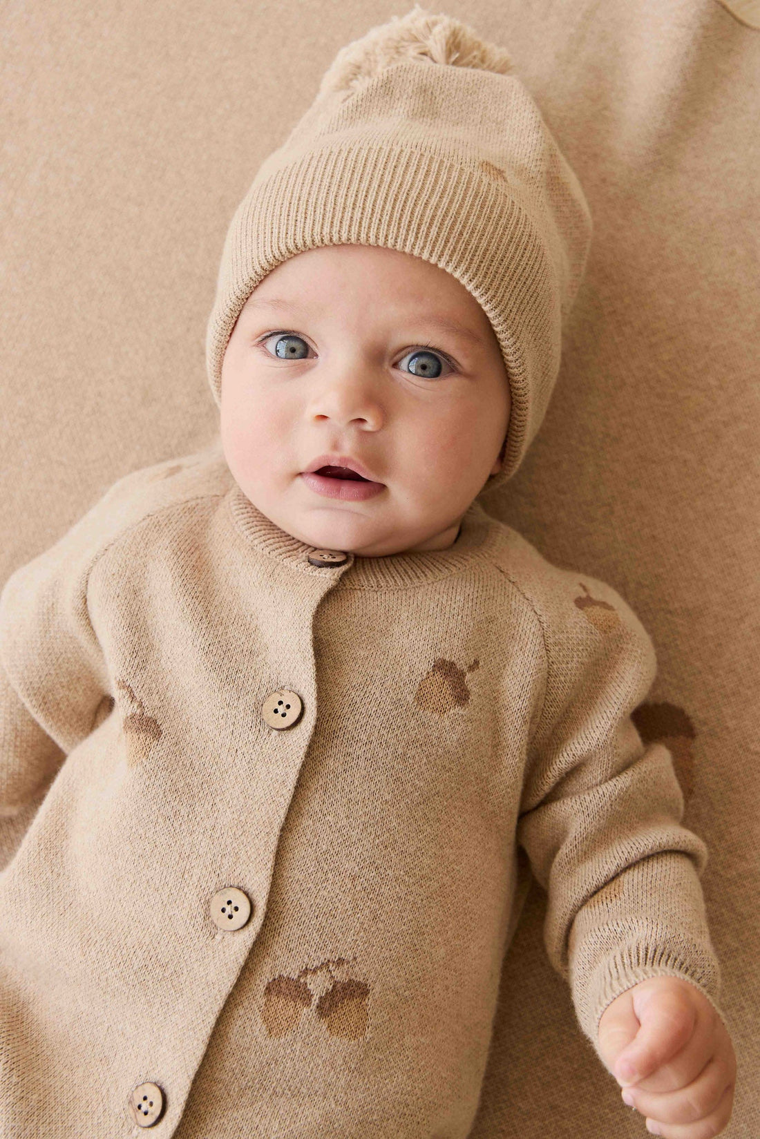 Baby wearing a beige knit outfit with a matching hat on a beige background