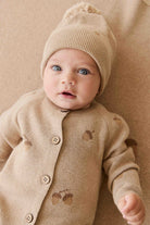 Baby wearing a beige knit outfit with a matching hat on a beige background