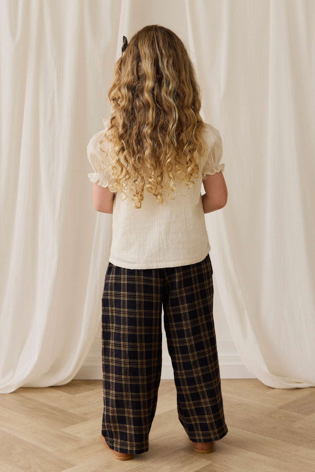 Child wearing a white top and plaid pants standing in front of white curtains.