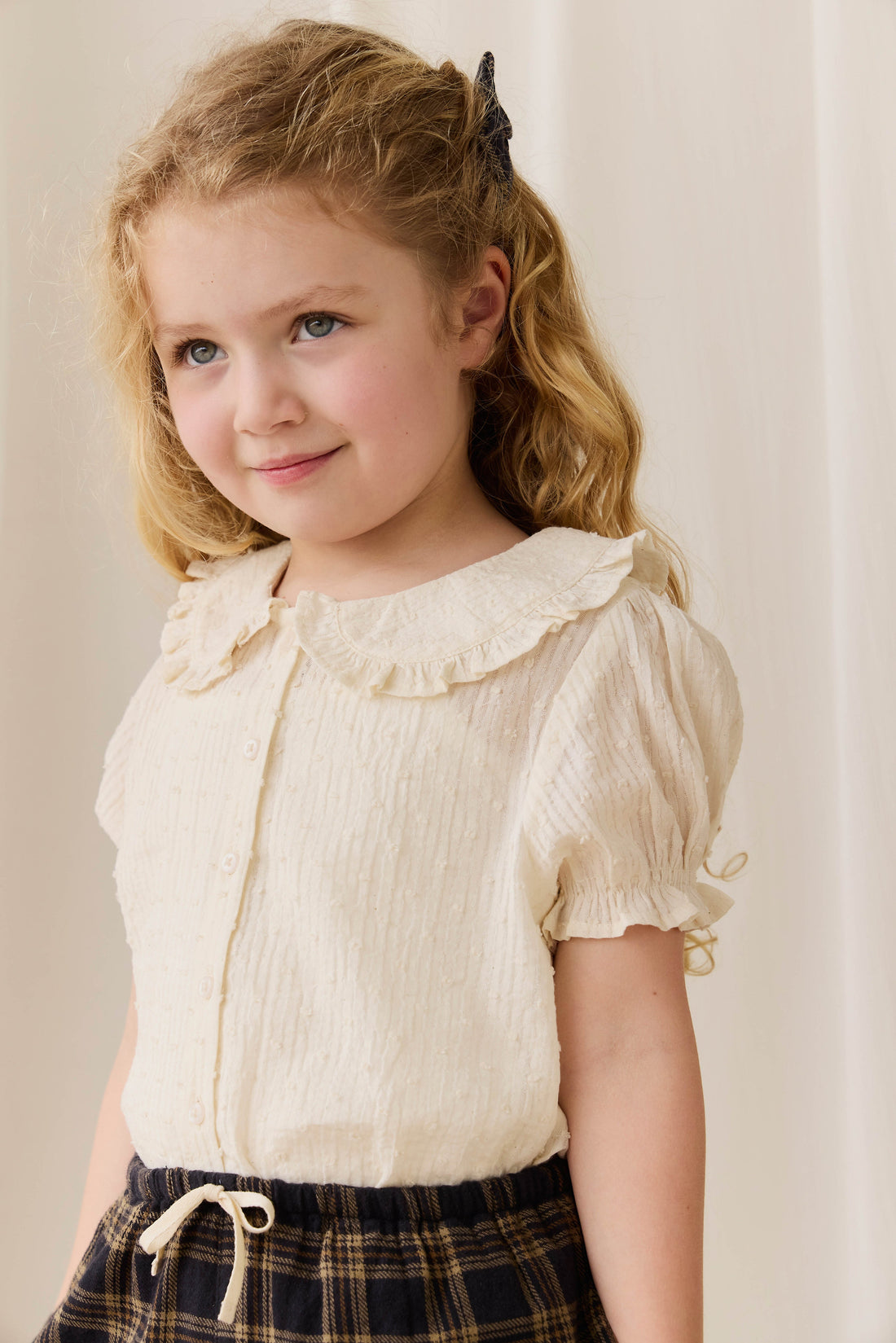 Young girl wearing a light-colored blouse with a ruffled collar against a plain background