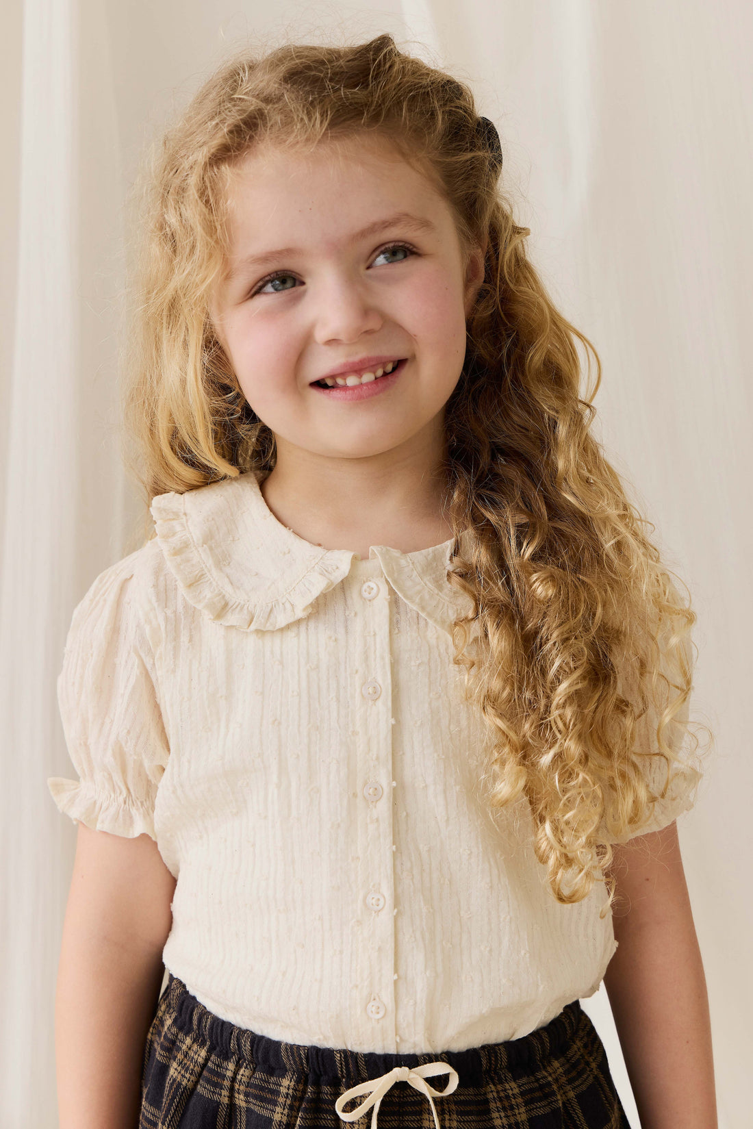 Young girl with curly hair wearing a white blouse and plaid skirt against a plain background