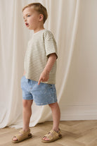 Child wearing a striped shirt and denim shorts standing against a white curtain.