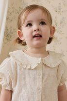 Young child wearing a white dress with a ruffled collar against a floral wallpaper background