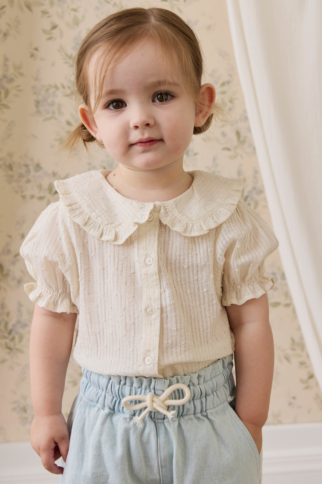 Young child wearing a white blouse with ruffled collar and light blue shorts.
