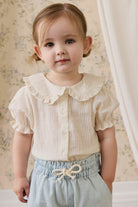 Young child wearing a white blouse with ruffled collar and light blue shorts.