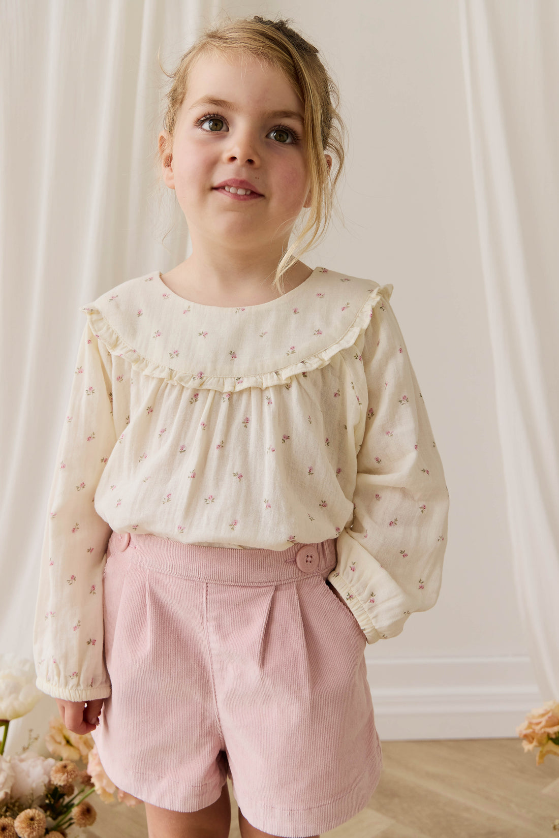 Young girl wearing a cream blouse with ruffled collar and pink shorts, standing against a white curtain.