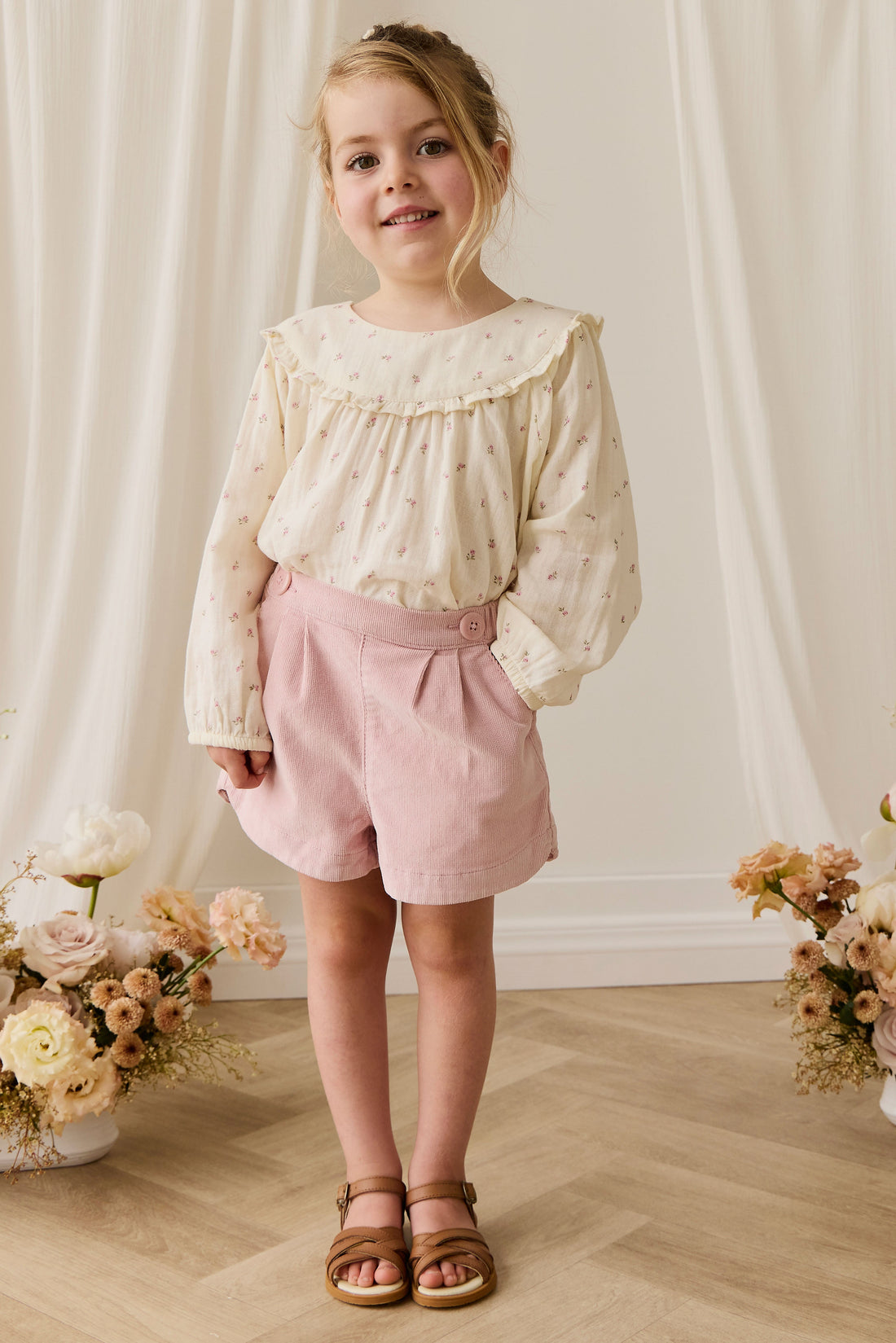 Young girl in a white blouse and pink shorts standing in front of a white curtain with floral decorations on either side.
