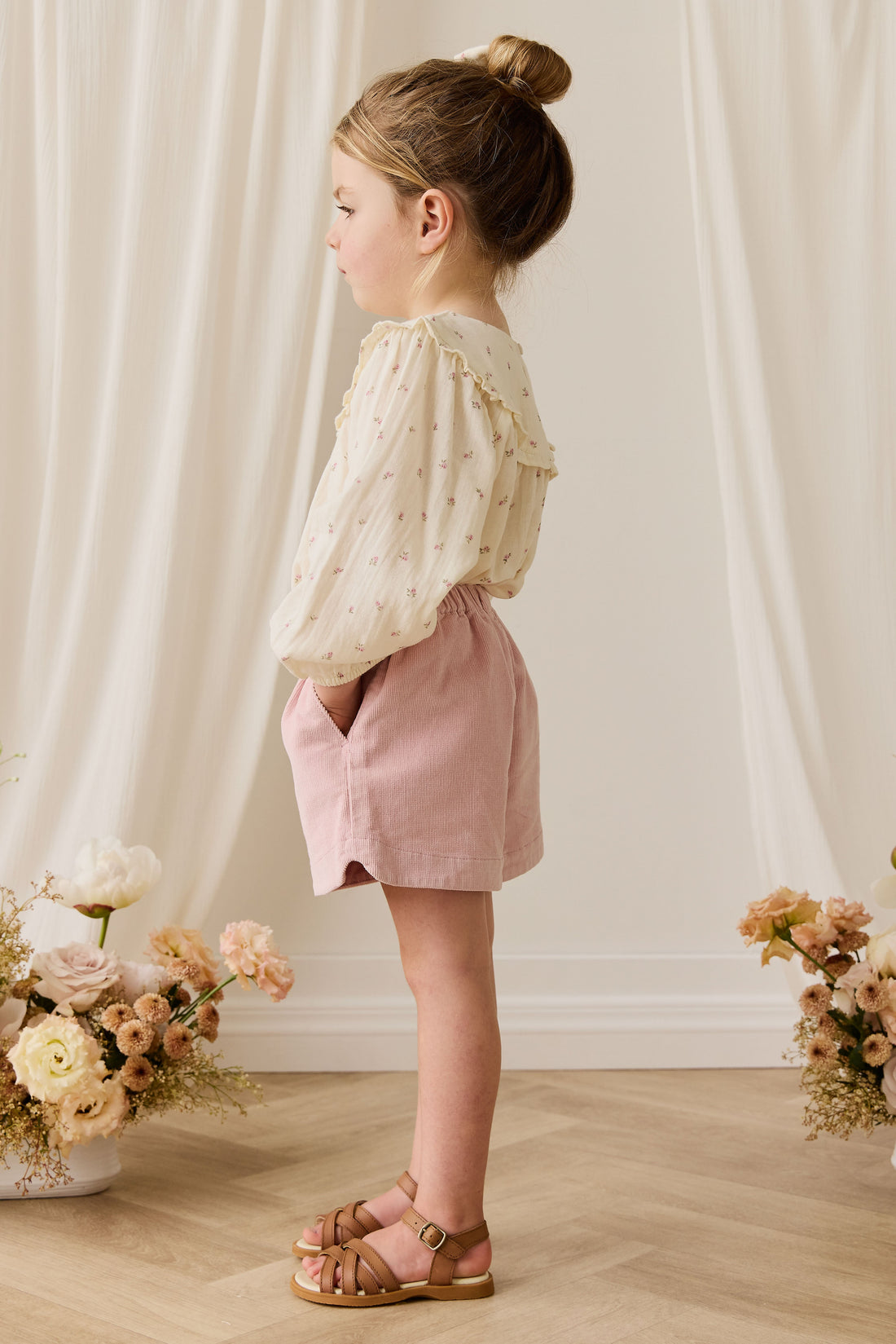 Young girl in a light-colored blouse and pink shorts standing in front of white curtains with floral arrangements on either side.