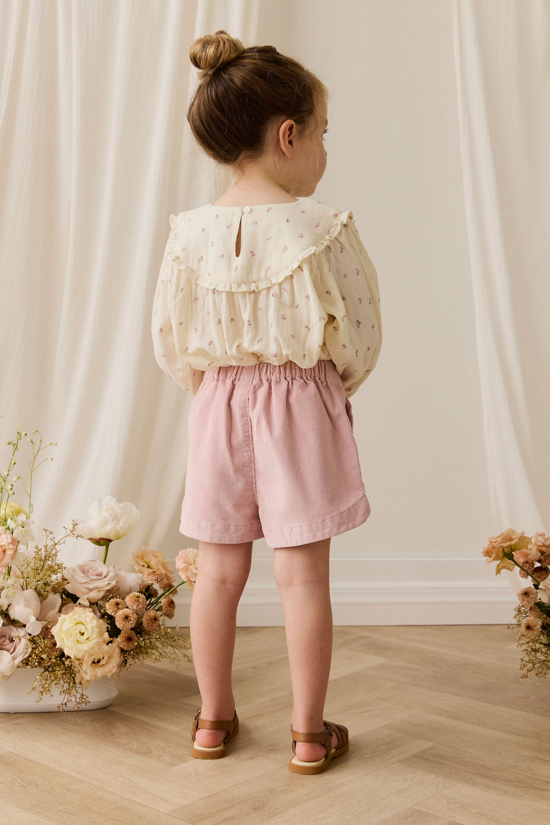 Young girl in a floral blouse and pink shorts standing in front of a white curtain with flowers on either side.