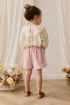 Young girl in a floral blouse and pink shorts standing in front of a white curtain with flowers on either side.