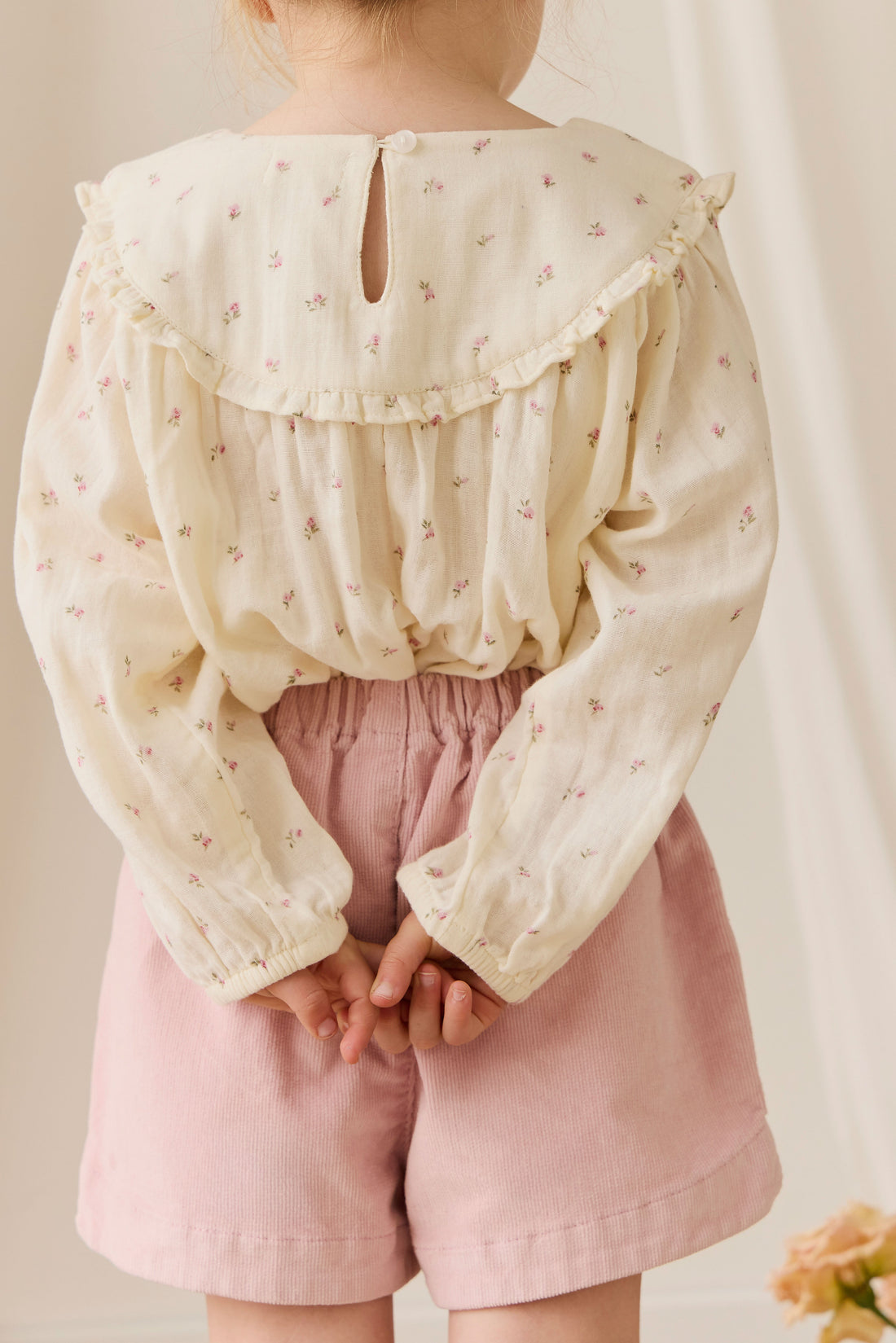 Child wearing a light-colored blouse with ruffled collar and pink shorts against a neutral background