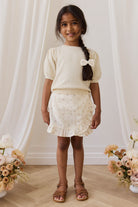 Young girl in a cream outfit with a bow standing between two flower arrangements.