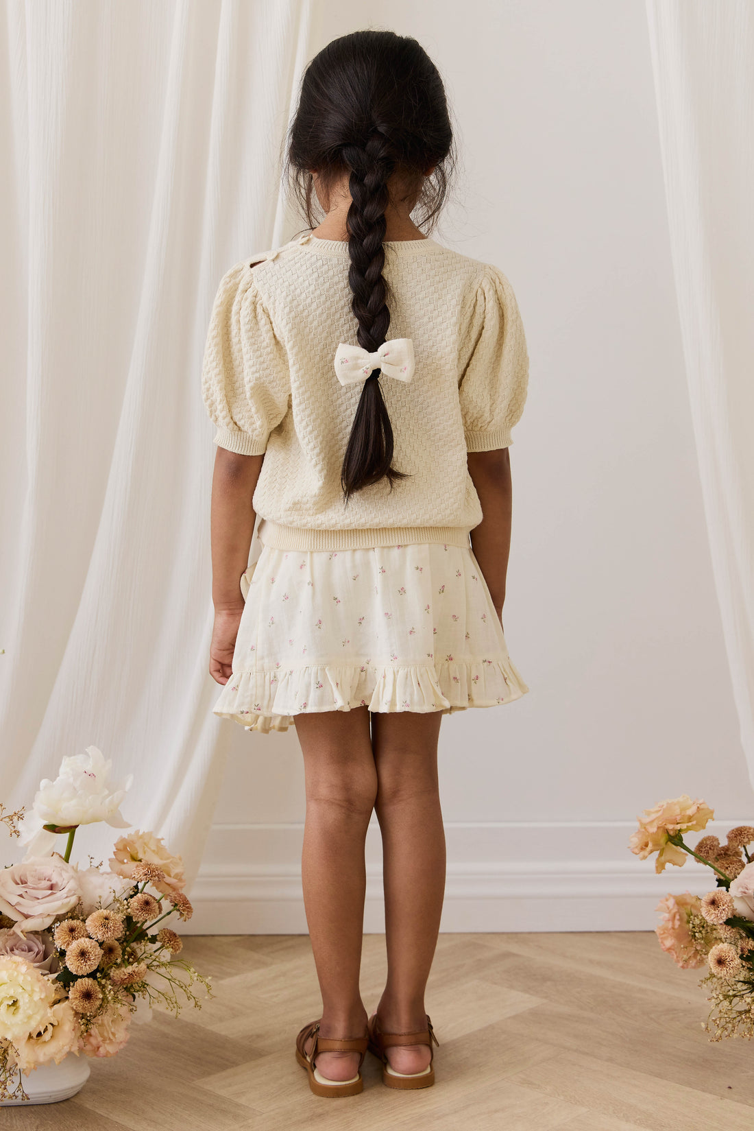 Young girl in a cream outfit with a bow, standing in front of white curtains with floral arrangements on either side.