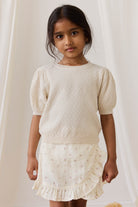 Young girl wearing a textured white top and ruffled skirt against a plain background