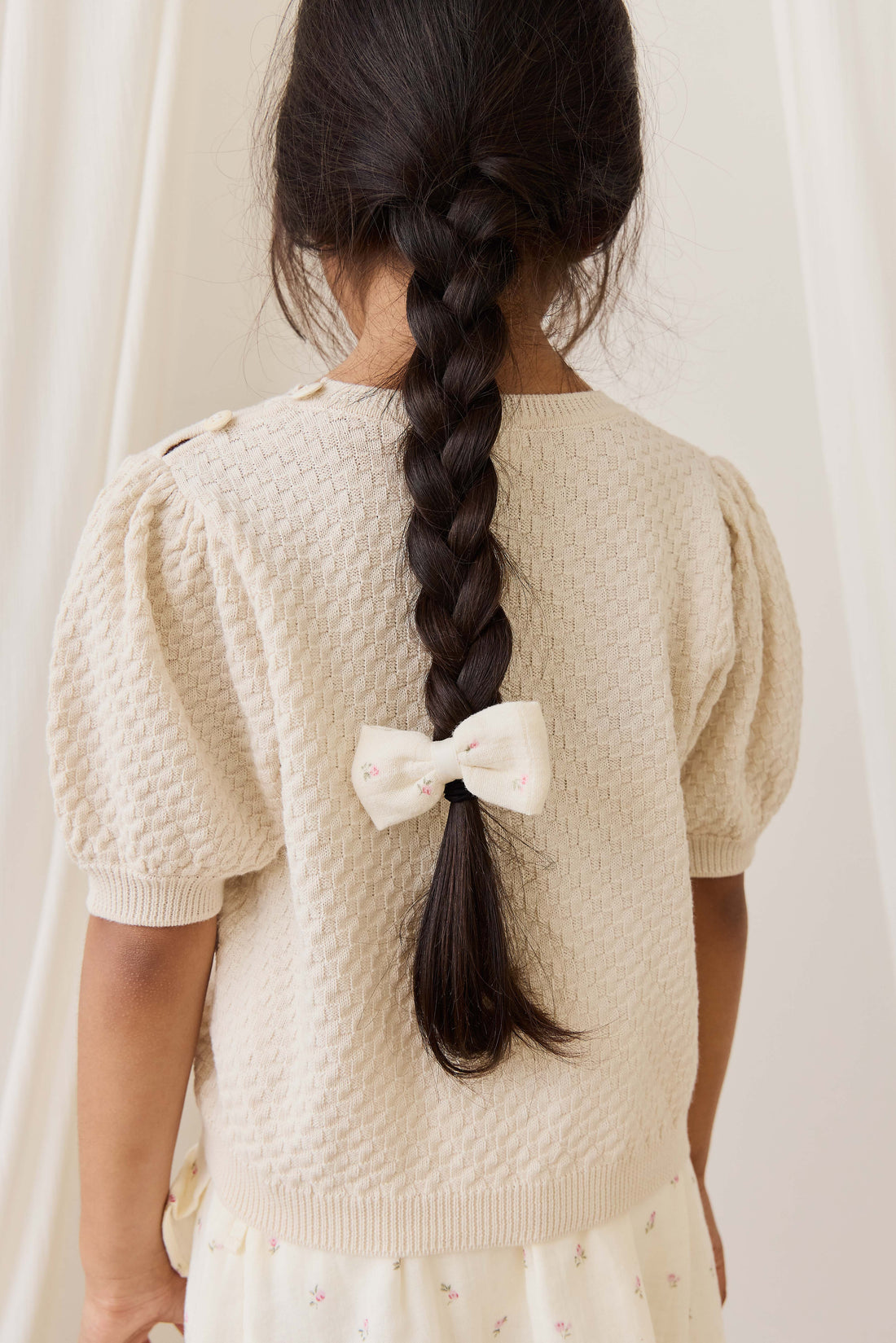 Child wearing a beige textured sweater with a white bow, standing against a white curtain.